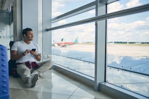 Happy smiling male sitting on the floor in waiting department in front of glass window at airport while looking to the plane in outdoors