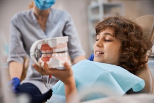 Neugieriges Kind, das ein Zahnmodell hält und aufmerksam betrachtet, während es mit einem Zahnarzt an seiner Seite im Zahnarztstuhl sitzt