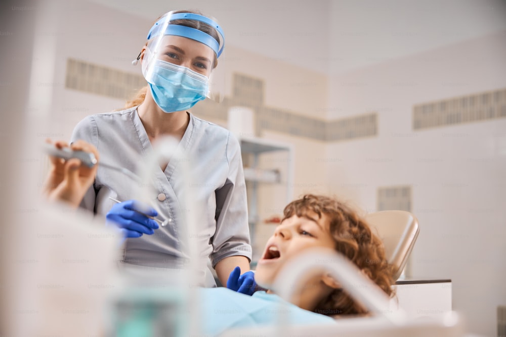 전문 여성 치과 의사는 마스크를 착용하고 아이와 약속하는 동안 구강 거울을 들고 있습니다.