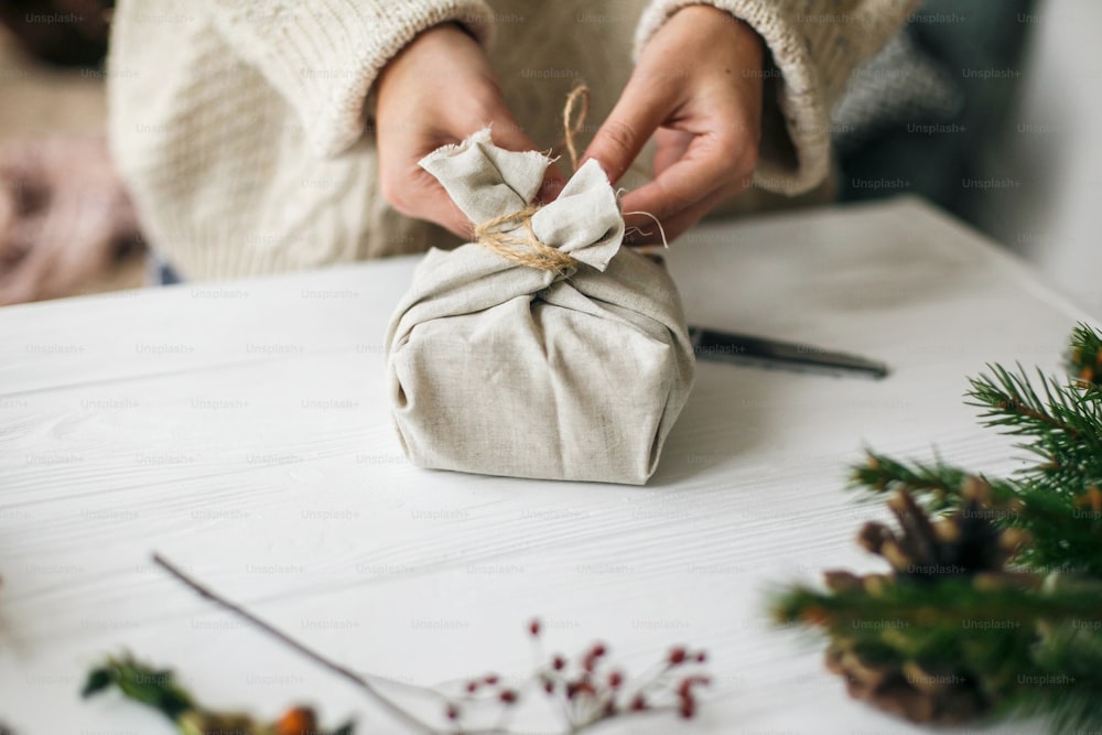 Woman in cozy sweater preparing plastic free christmas present. Female hands wrapping stylish christmas gift in linen fabric on white rustic table with green branch, pine cones, scissors and twine.