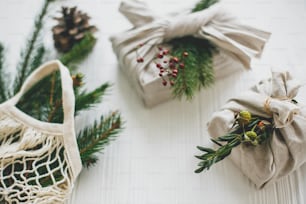 Stylish christmas gifts wrapped in linen fabric with green branch and reusable shopping bag with green spruce on rustic wooden background. Zero waste christmas holidays. Sustainable lifestyle