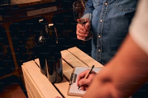 Cropped photo of unrecognized male sommelier standing in the cellar with a glass of red wine while his assistant making notes