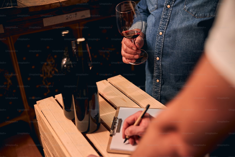 Cropped photo of unrecognized male sommelier standing in the cellar with a glass of red wine while his assistant making notes