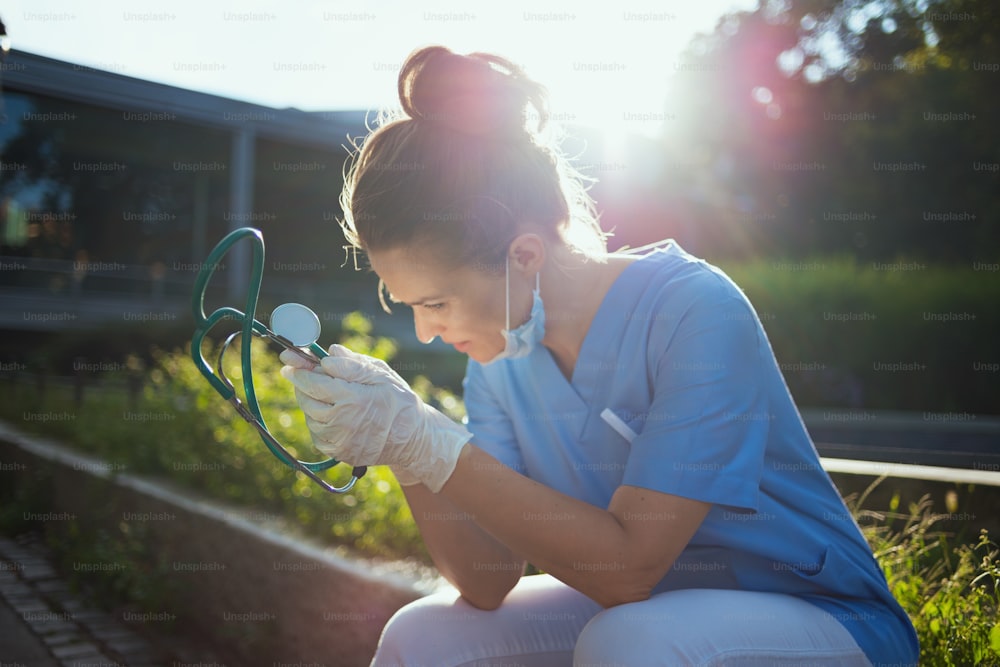 coronavirus pandemic. sad modern physician woman in uniform with stethoscope and medical mask sitting outside near clinic.