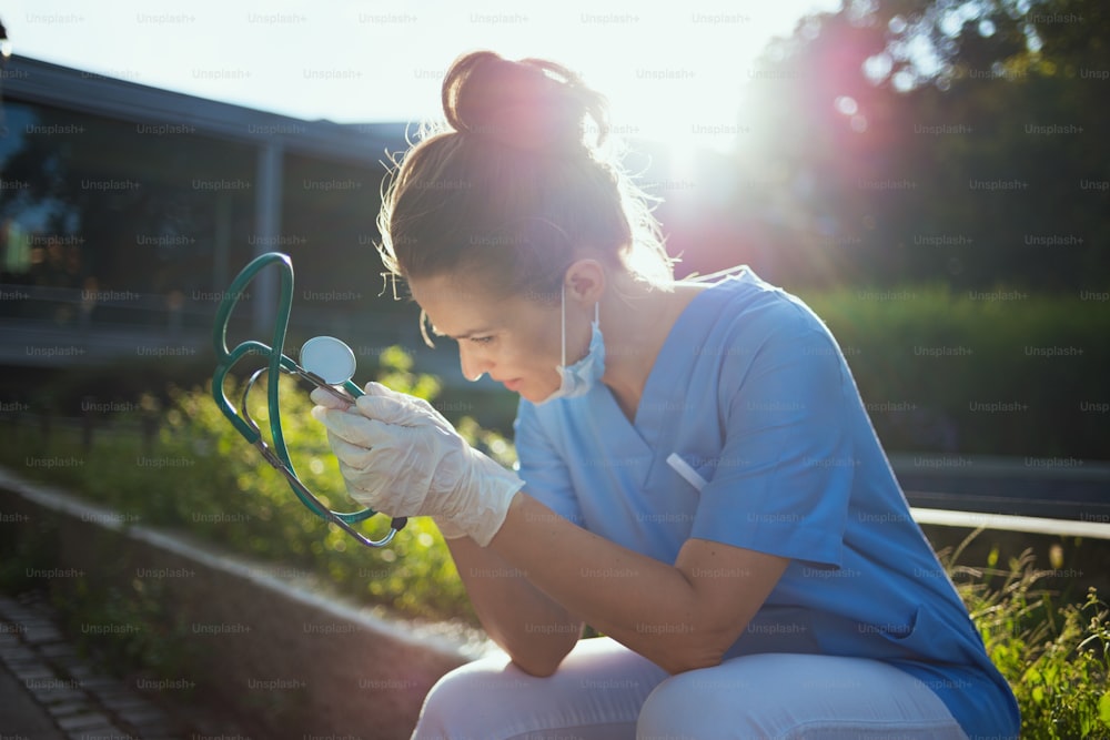 코로나 바이러스 전염병. 청진기와 의료용 마스크를 쓴 제복을 입은 슬픈 현대 의사 여성이 병원 근처에 앉아 있다.