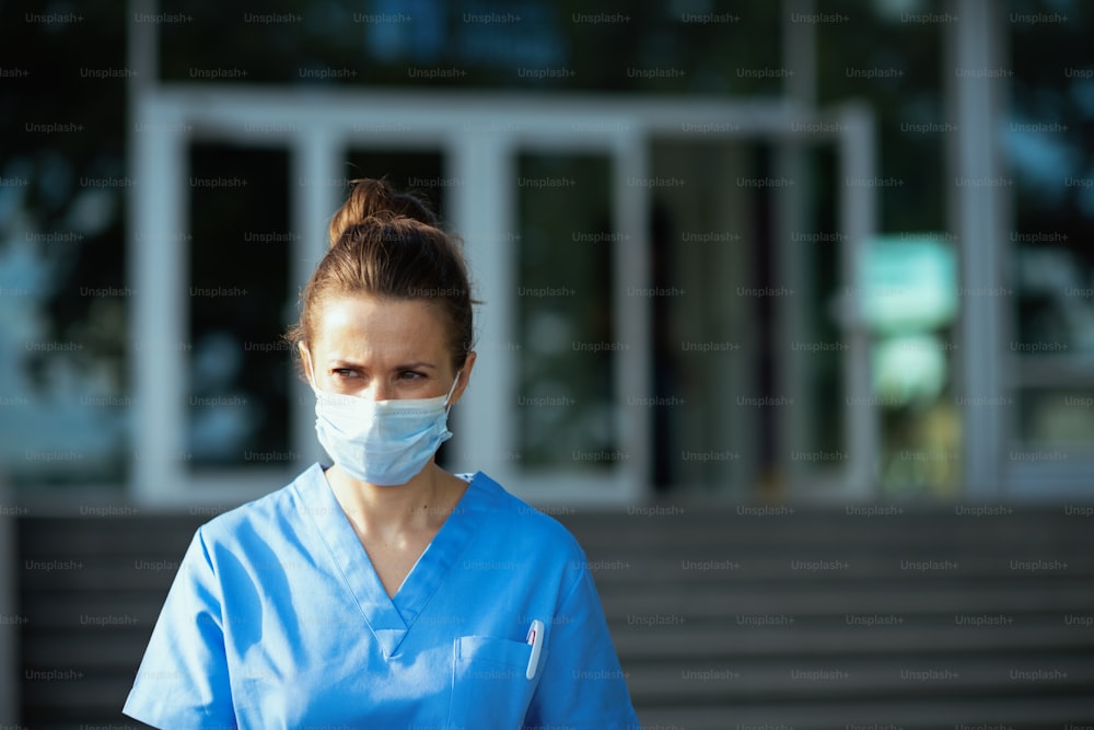 Pandemia di COVID-19. donna medica moderna infelice in uniforme con maschera medica all'aperto vicino all'ospedale.