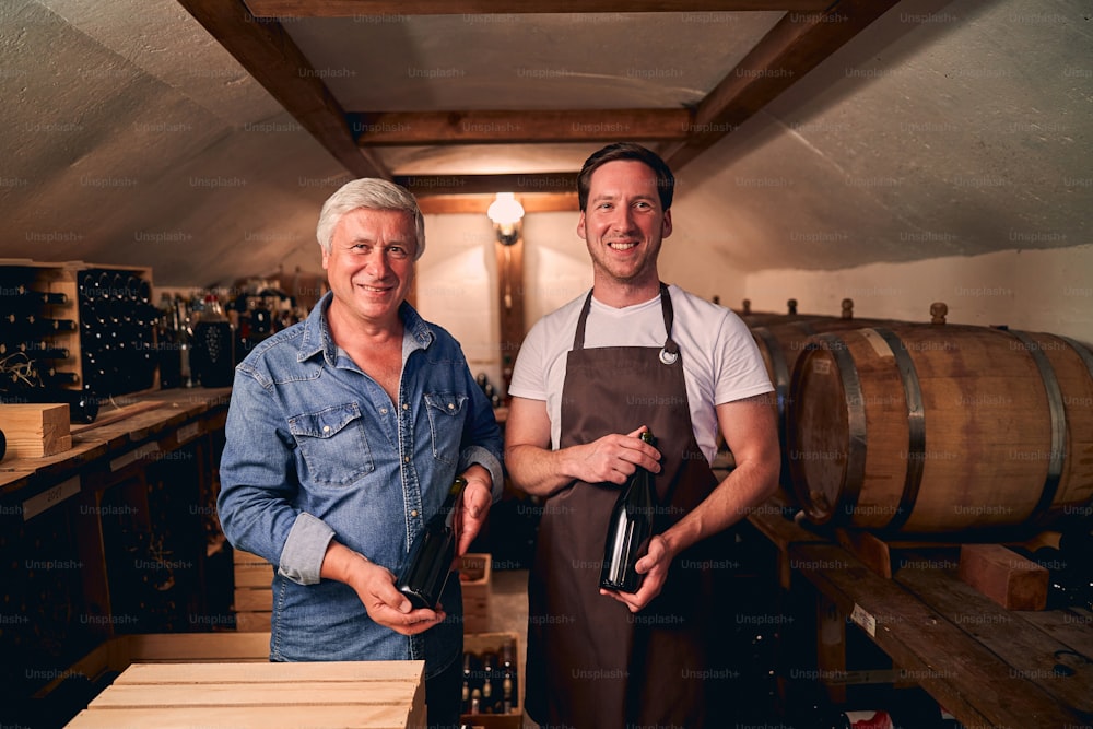 Cheerful Caucasian males smiling happily while standing in a cellar with unlabeled bottles of wine. Barrels on the background
