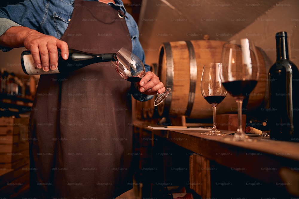 Cropped photo of a male sommelier in a brown apron standing in the cellar and pouring wine into a glass