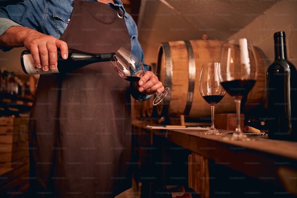 Cropped photo of a male sommelier in a brown apron standing in the cellar and pouring wine into a glass