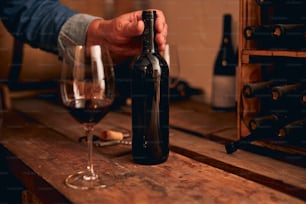 ボトルネックに手を当てる正体不明の男。木製のテーブルの上に置かれた赤ワインのグラス