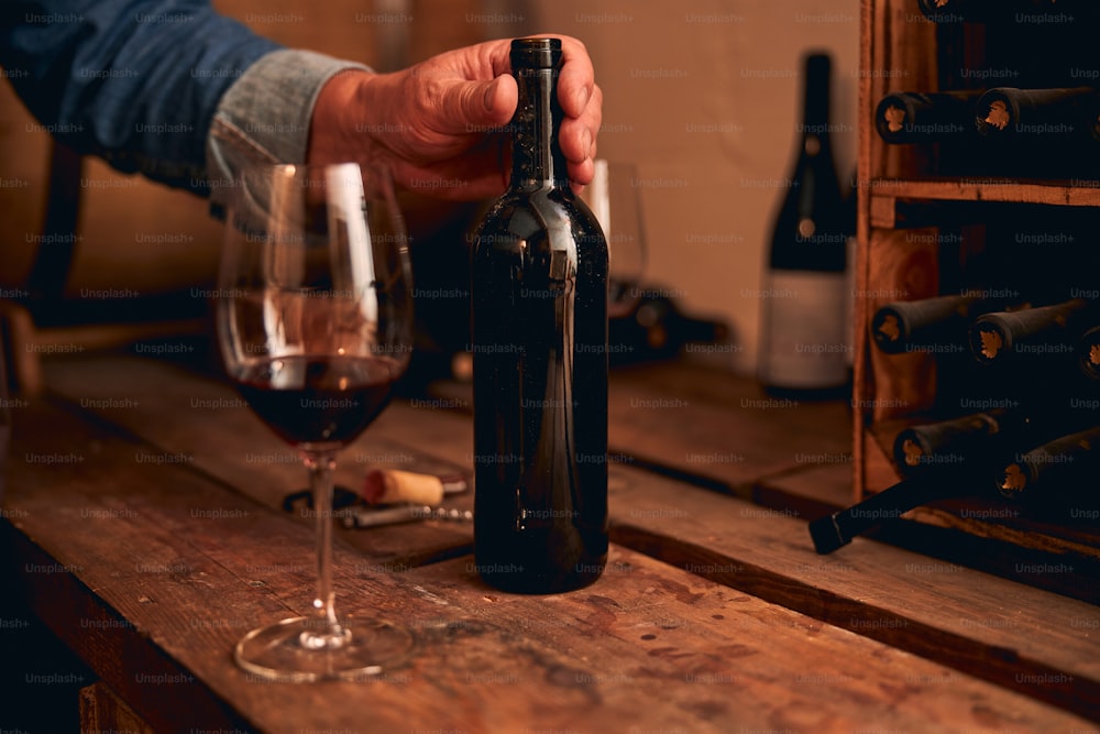 Unerkannter Mann legt seine Hand auf einen Flaschenhals. Glas Rotwein in der Nähe auf Holztisch