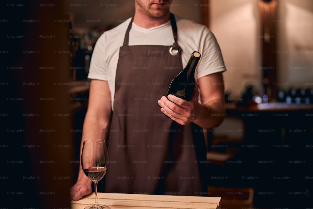Photo recadrée d’un homme de race blanche vêtu d’un tablier brun debout avec une bouteille bouchée. Un verre de vin devant lui sur une table