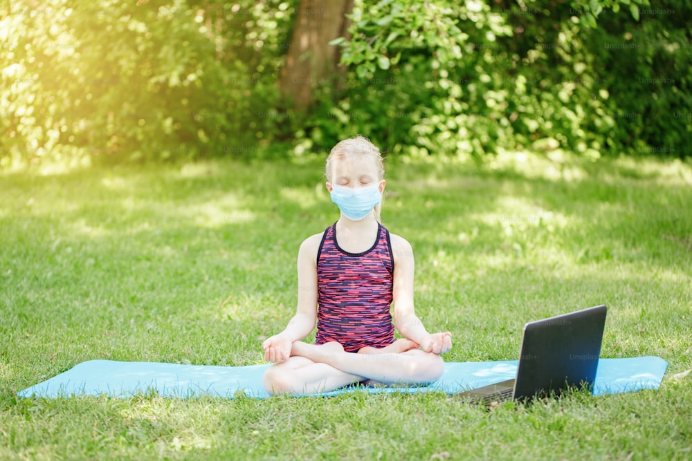 Kaukasisches Mädchen in Gesichtsmaske macht Sport Workout Outdoor online. Video Yoga im Internet. Kind lernt Training im Hinterhof zu Hause mit Laptop. Neue Normalität. Soziale Distanz beim Coronavirus.