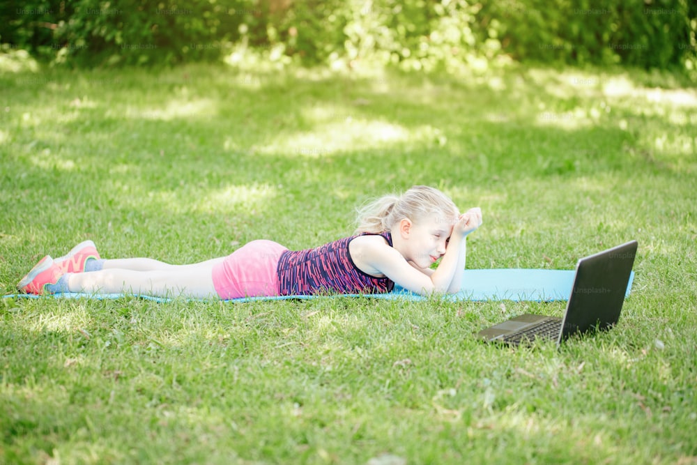 Bored tired girl child doing sport workout outdoor online. Video yoga class on Internet. Funny sleepy kid training at home backyard park with laptop. New normal. Social distance at coronavirus.