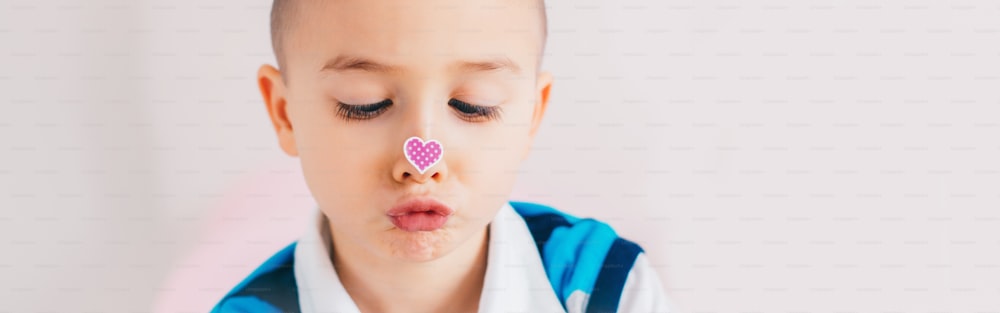 Cross-eyed kid looking at purple violet heart sticker on nose. Funny hilarious white Caucasian cute adorable child boy making faces. Valentine Day holiday concept. Web banner header for website.