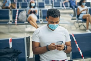 Junger indischer Geschäftsmann sitzt und benutzt Smartphone im Wartebereich des internationalen Flughafens
