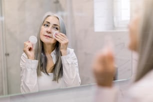 Skin care and hygiene concept. Charming senior gray haired lady using a cotton pad with micellar water for removing make up from face, standing in bathroom and looking in the mirror.