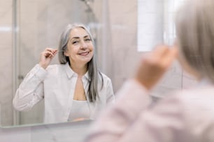Portrait of pretty senior gray haired woman looking to mirror, cleansing her ears with a cotton swab in modern light bathroom at home. Beauty, hygiene and people concept.