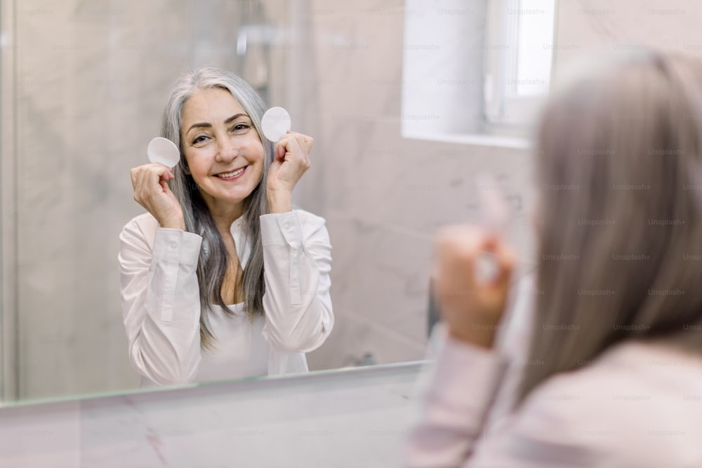 Smiling pretty mature woman with beautiful long gray hair, performing her skincare routine, posing in home light bathroom in front of the mirror with cotton pads.