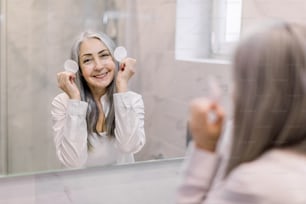 Lächelnde hübsche reife Frau mit schönen langen grauen Haaren, die ihre Hautpflegeroutine durchführt und im hellen Badezimmer vor dem Spiegel mit Wattepads posiert.
