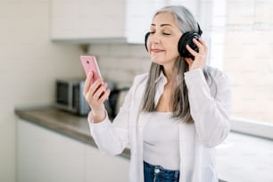 Enjoying music and having fun in the kitchen. Cheerful old gray haired woman is listening to music from earphones and smiling, or havinf video call with best friend. Elderly people and technologies.