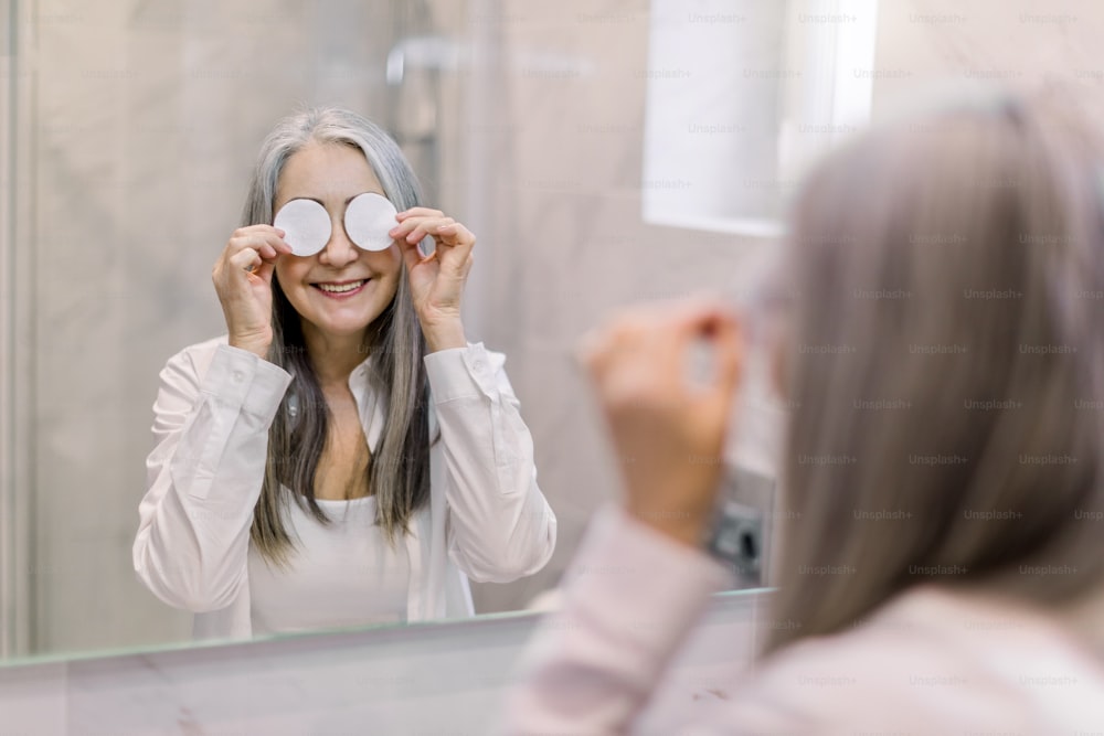 Face skin care and beauty procedures. Portrait of positive funny elderly gray haired woman, standing in the bathroom in front of mirror and covering her eyes with cotton pads.