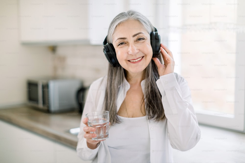 Retrato de una abuela madura feliz y sonriente con el pelo largo y gris, de pie en una cocina moderna y luminosa con un vaso de agua, escuchando la música o el audiolibro con auriculares