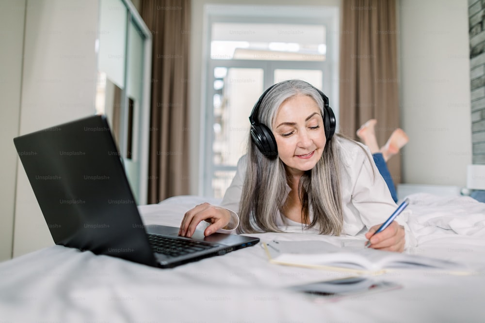 Schöne Senior konzentrierte Geschäftsfrau in Kopfhörern, schreibt Notizen, während Sie Video-Online-Webinar ansehen, lernen oder arbeiten am Laptop zu Hause, auf dem Bett liegend. Fern-E-Learning-Konferenz.
