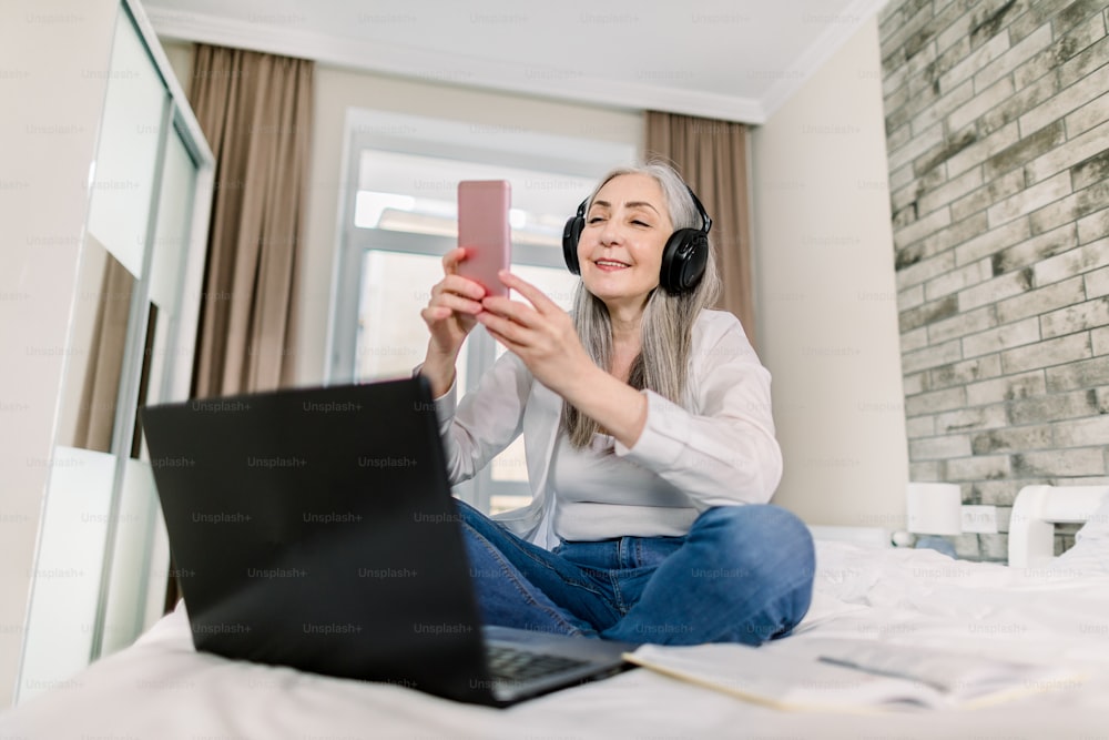 Senior Peope und Technologien Konzept. Hübsch lächelnde pensionierte grauhaarige Frau mit Kopfhörern, die auf dem Bett sitzt und ihr Smartphone für Videochat oder das Tippen von Nachrichten benutzt, während sie am Laptop arbeitet.