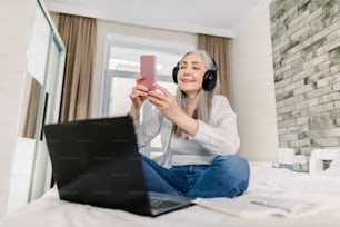 Senior Peope und Technologien Konzept. Hübsch lächelnde pensionierte grauhaarige Frau mit Kopfhörern, die auf dem Bett sitzt und ihr Smartphone für Videochat oder das Tippen von Nachrichten benutzt, während sie am Laptop arbeitet.