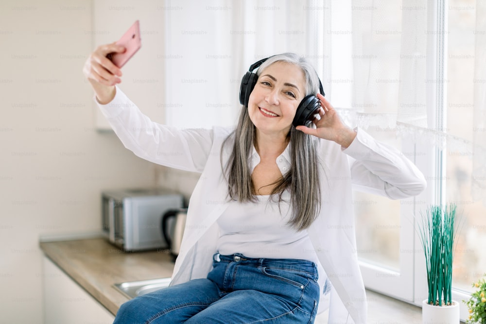 Modern senior people and technologies concept. Smiling retired happy woman using smartphone and headphones for listening music and making video calls or photo, in the kitchen at home