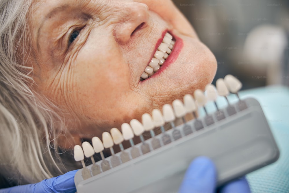 Portrait de vue latérale d’une femme âgée souriante assise dans le cabinet dentaire tandis que le médecin en gant bleu tenant un membre artificiel en porcelaine