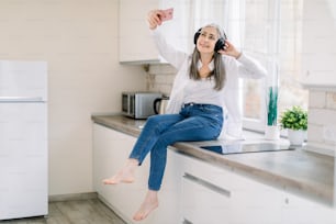 People at home concept. Happy Caucasian elderly woman with headphones holding mobile phone for making photo or video call, while sitting on kitchen countertop in modern kitchen.