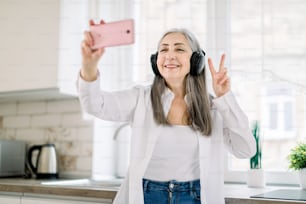 Portrait d’une dame âgée positive, heureuse et souriante avec de longs cheveux gris, avec des écouteurs, s’amusant dans la cuisine à la maison, écoutant de la musique et faisant une photo sur le téléphone portable faisant un signe de victoire.