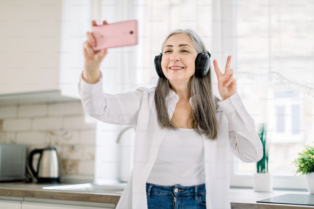 Portrait of positive, happy, smiling senior lady with long gray hair, with headphones, having fun in kitchen at home, listening to music and making photo on cell phone gesturing victory sign.