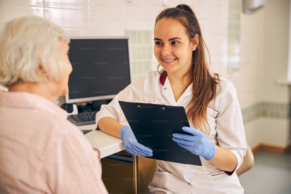 Confident doctor cosmetologist working with documents in modern cosmetic office while talking with patient
