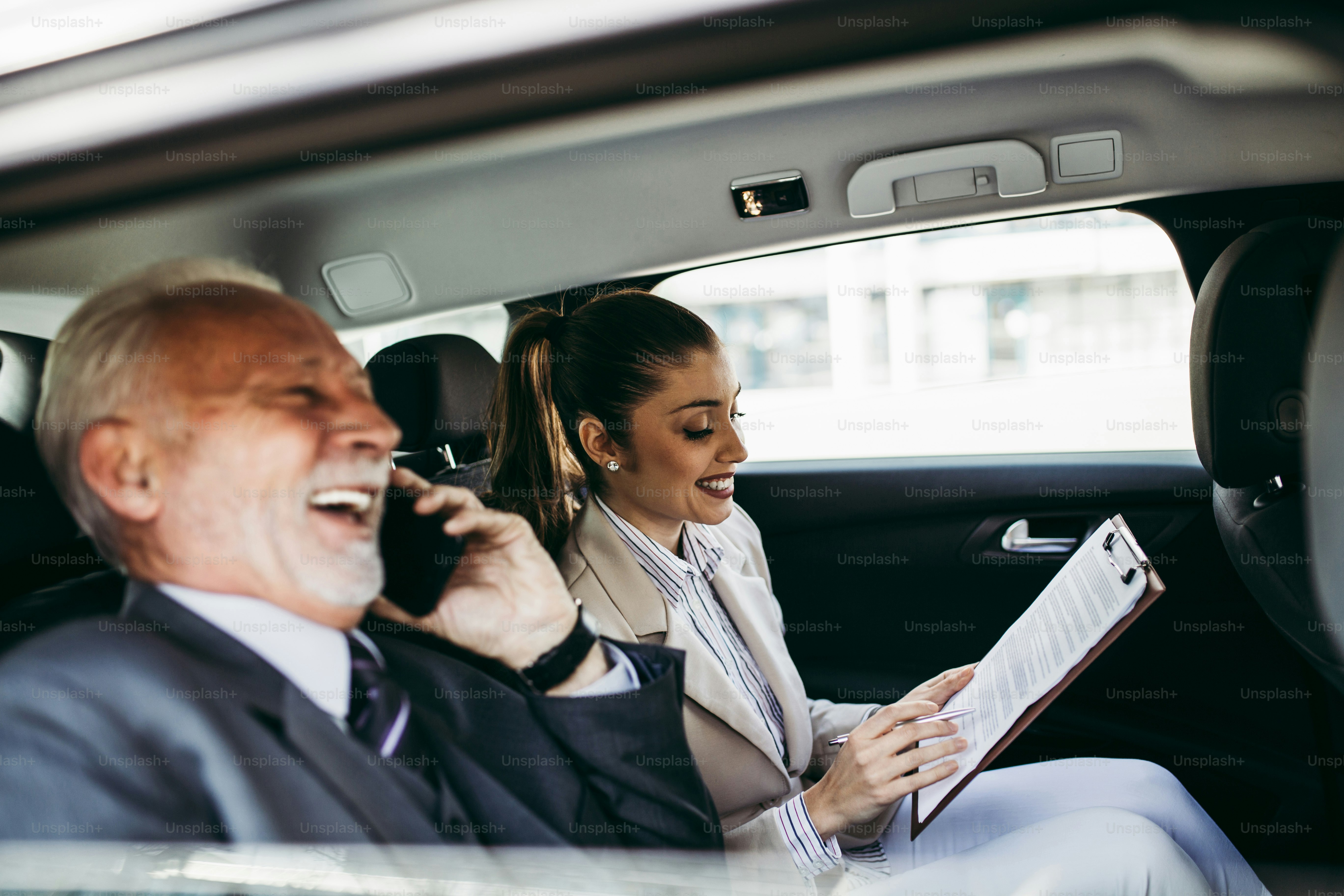 Good looking senior business man and his young woman colleague or coworker sitting on backseat in luxury car. They talking, smiling and using laptop and smart phones. Transportation in corporate business concept.