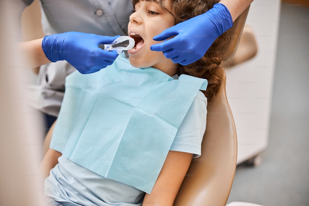 Beschnittenes Foto eines Kindes, das seinen Mund öffnet, um einen Zahngummidamm von einem Zahnarzt einsetzen zu lassen