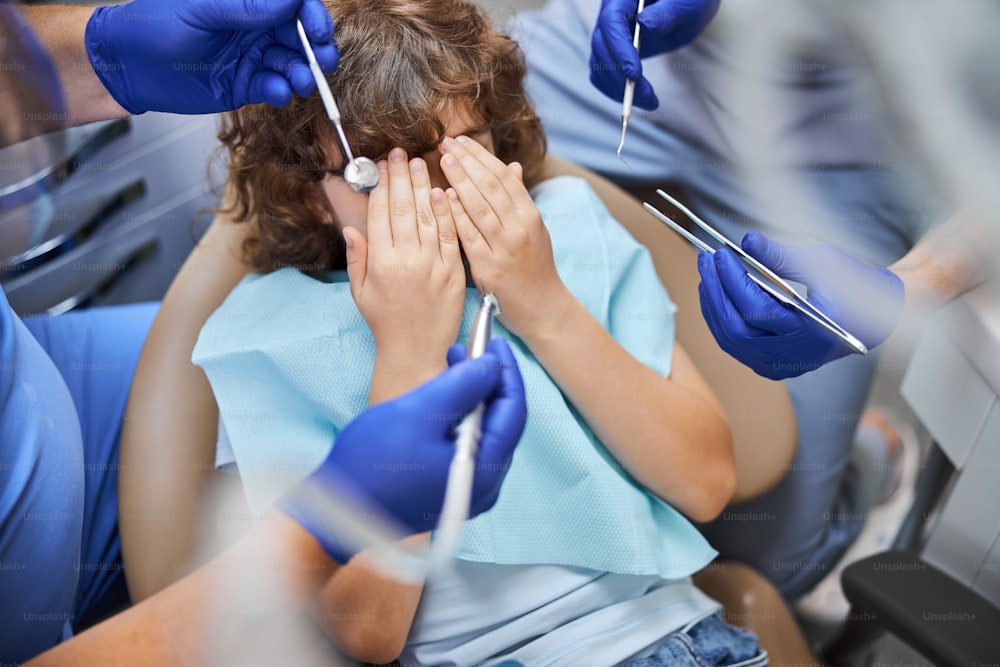Scared brown-haired child covering his face with hands while doctors holding dental tools all around him