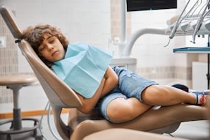 Foto de un niño moreno rizado con un babero sanitario durmiendo la siesta pacíficamente en una silla en el laboratorio del dentista