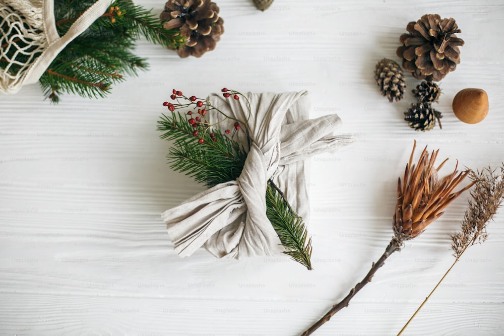 Pose plate de Noël zéro déchet. Cadeau de Noël élégant enveloppé dans un tissu de lin, décoré d’une branche verte naturelle sur fond de table rustique blanc avec des pommes de pin et des herbes.