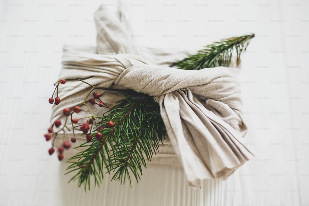 Elegante regalo di Natale avvolto in tessuto di lino e decorato con ramo di abete naturale e bacche rosse su sfondo bianco rustico. Vacanze di Natale a rifiuti zero.