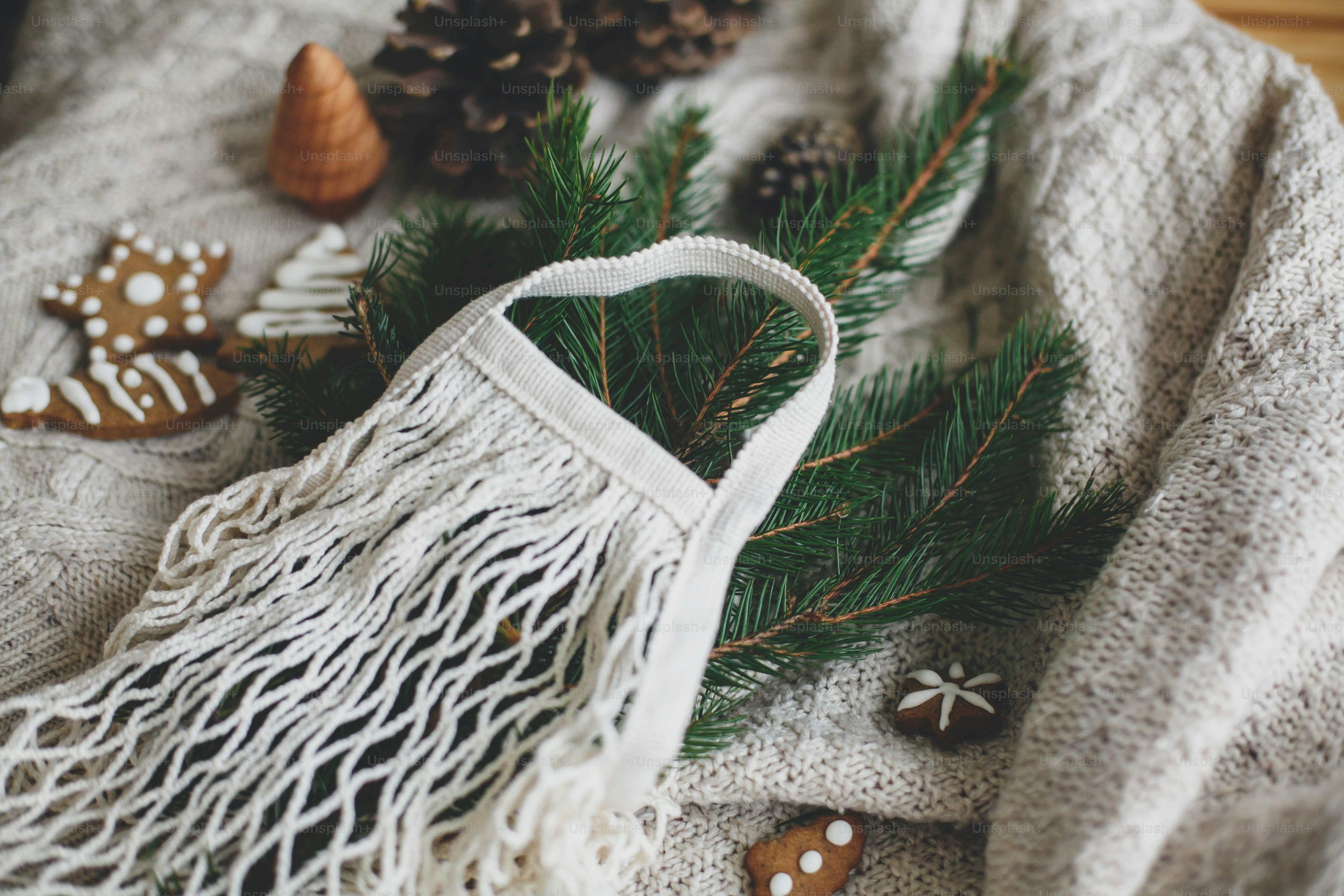 Reusable shopping bag and wooden winter decorations, plastic free holidays. Net cotton bag with green spruce branches, gingerbread christmas cookies and pine cones on white knitted sweater.