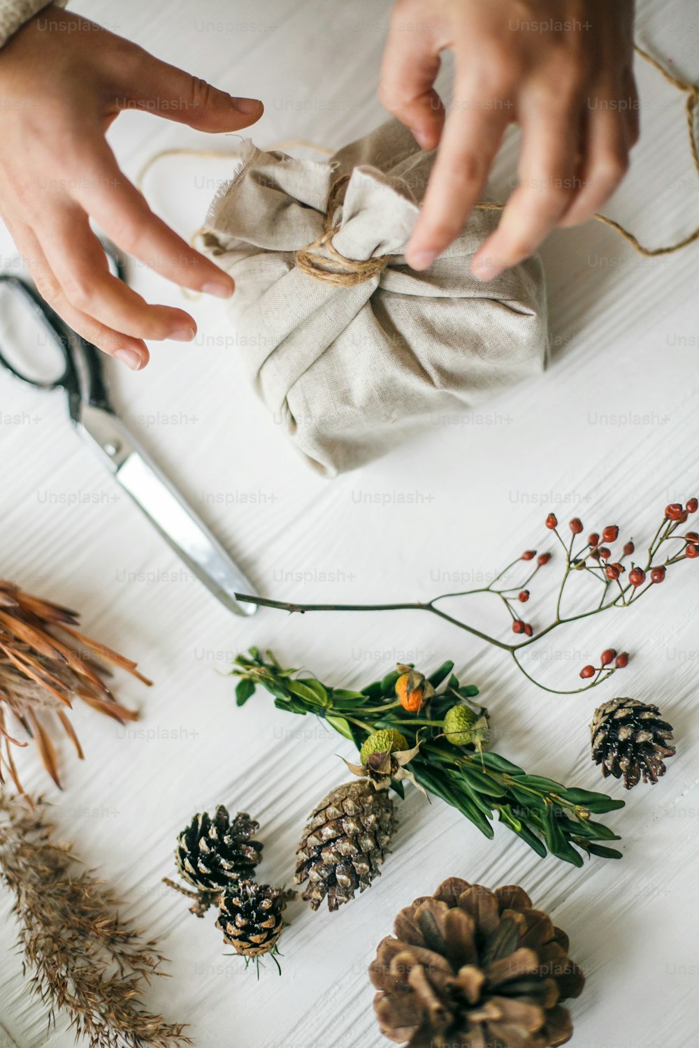 Hands wrapping stylish christmas gift in linen fabric on white rustic table with fir, pine cones, scissors, twine. Female preparing plastic free christmas present, zero waste holidays.