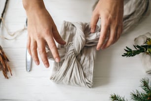 Plastic free holidays, zero waste Christmas. Hands wrapping christmas gift in linen fabric in furoshiki style on white rustic table with natural fir, herb, pine cones and scissors.