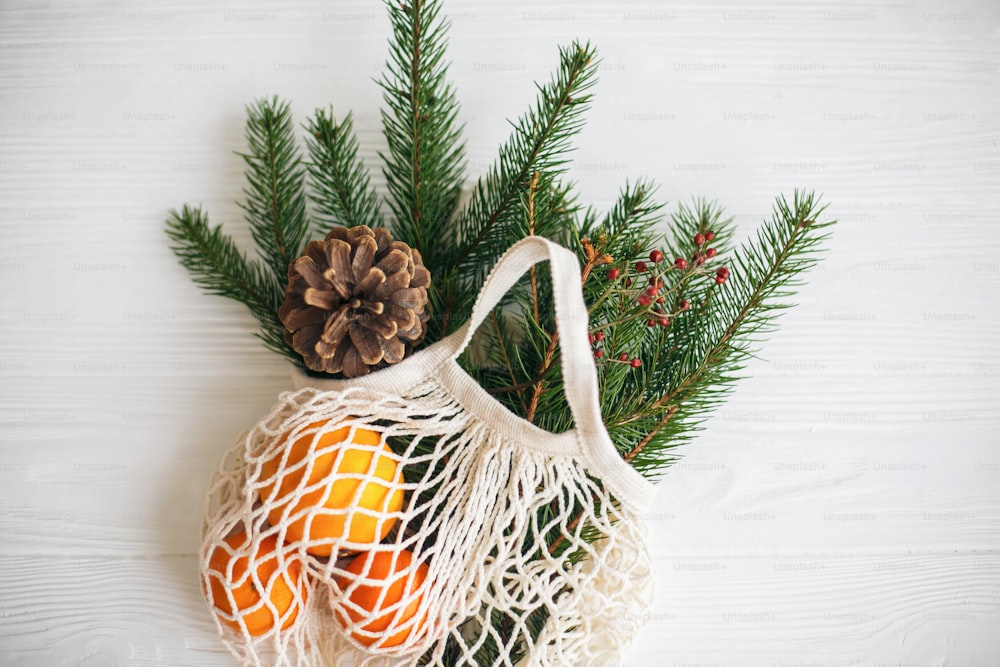 흰색 소박한 배경에 녹색 가문비나무 가지, 오렌지, 솔방울이 있는 재사용 가능한 면 가방. 겨울 장식, 제로 폐기물 휴일과 그물 쇼핑백. 상위 뷰