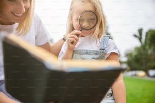 Happy cheerful mother with beautiful daughter studying material of book while sitting in the outdoors