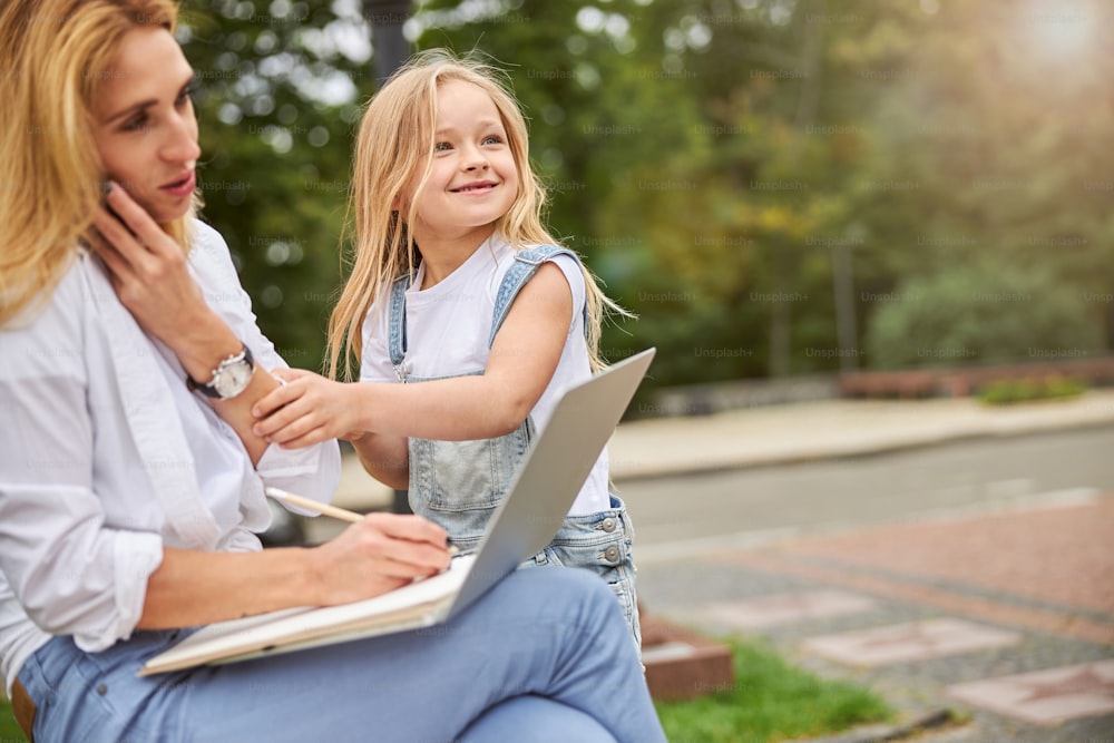 Pretty blonde mother and cute daughter working on a laptop in the park on a background of green trees