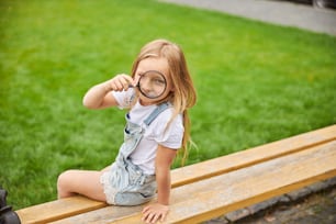 Charming girl holding magnifying glass while sitting on the wooden bench in the city park