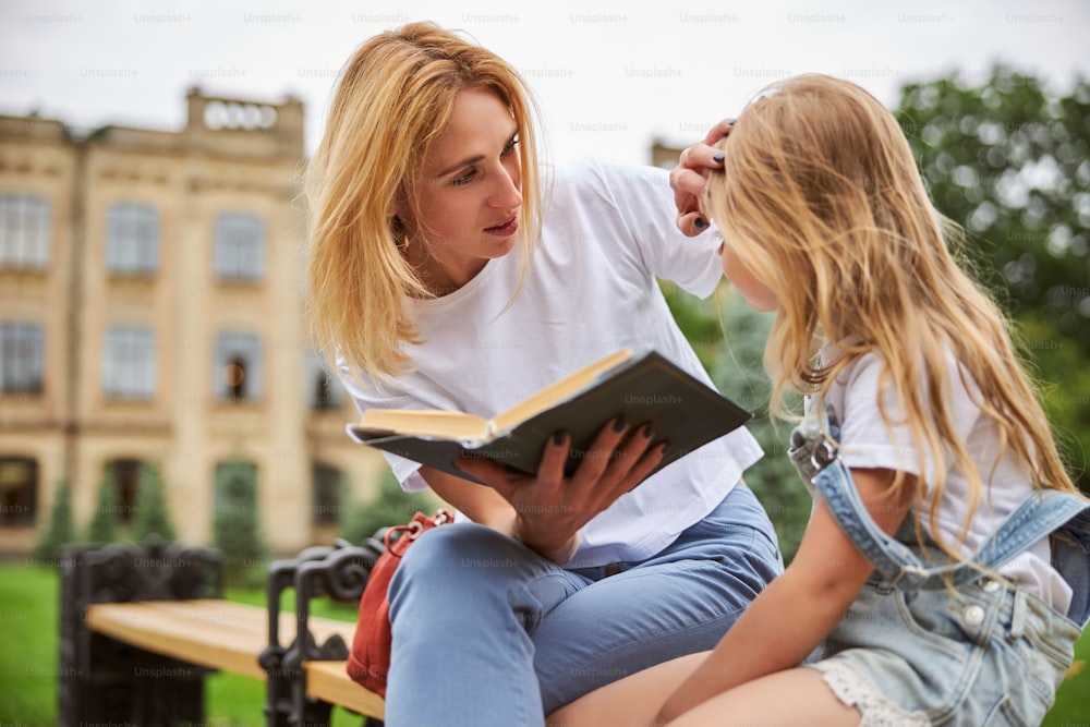 Cheerful blonde female reading a fairytale to her little daughter while spending time in the outdoors